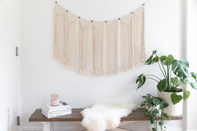 Macrame wall hanging over wooden bench with books stacked and houseplants in white pots